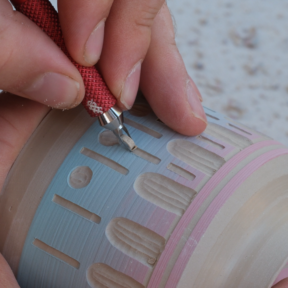 A close-up of hands holding a cylindrical ceramic vessel while using a red textured ceramic sculpting tool to carve decorative patterns into the surface.