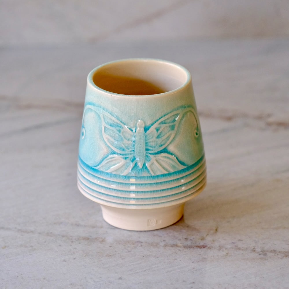 A small ceramic cup with a light blue butterfly design and a cream-colored base, set on a light gray marble surface.