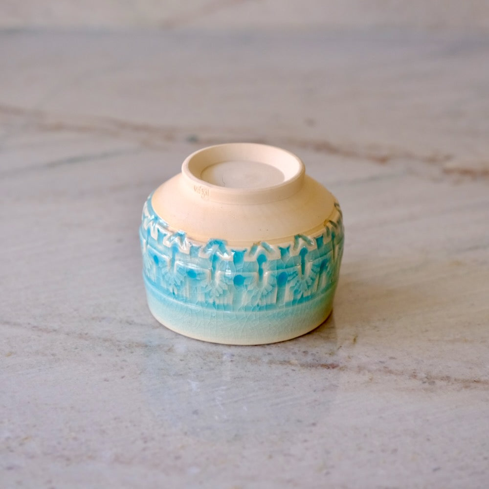 A small handmade ceramic cup with a cream-colored interior and exterior, featuring a textured turquoise blue pattern around the middle, resting on a light gray marble surface.