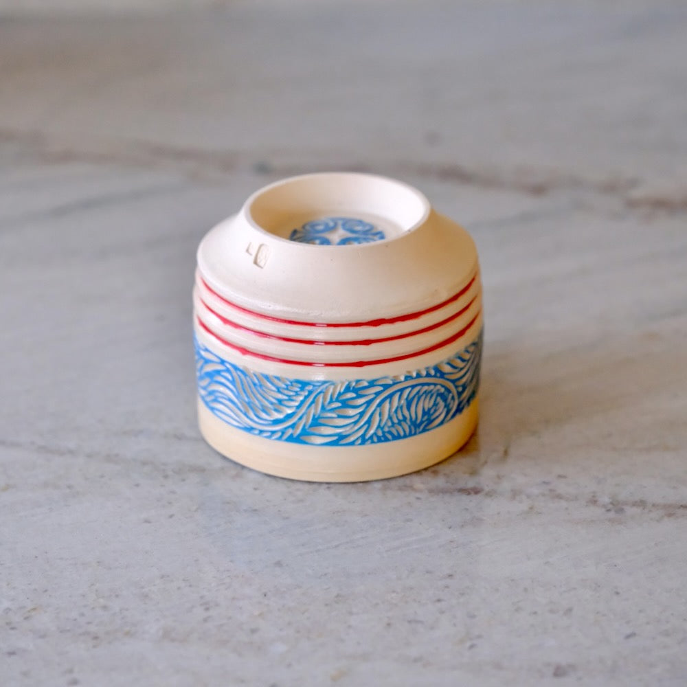 A small ceramic cup or candle holder with a cream-colored glaze, featuring a band of blue wave patterns and red horizontal stripes, set on a light gray marble surface.
