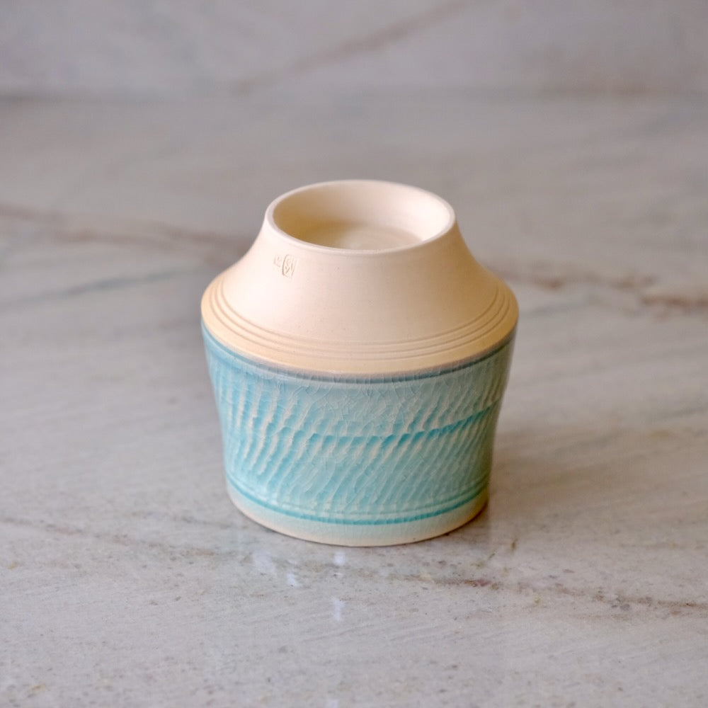 A small ceramic cup with a cream-colored unglazed top and a light blue glazed lower body featuring a textured wavy pattern, set on a light marble surface.
