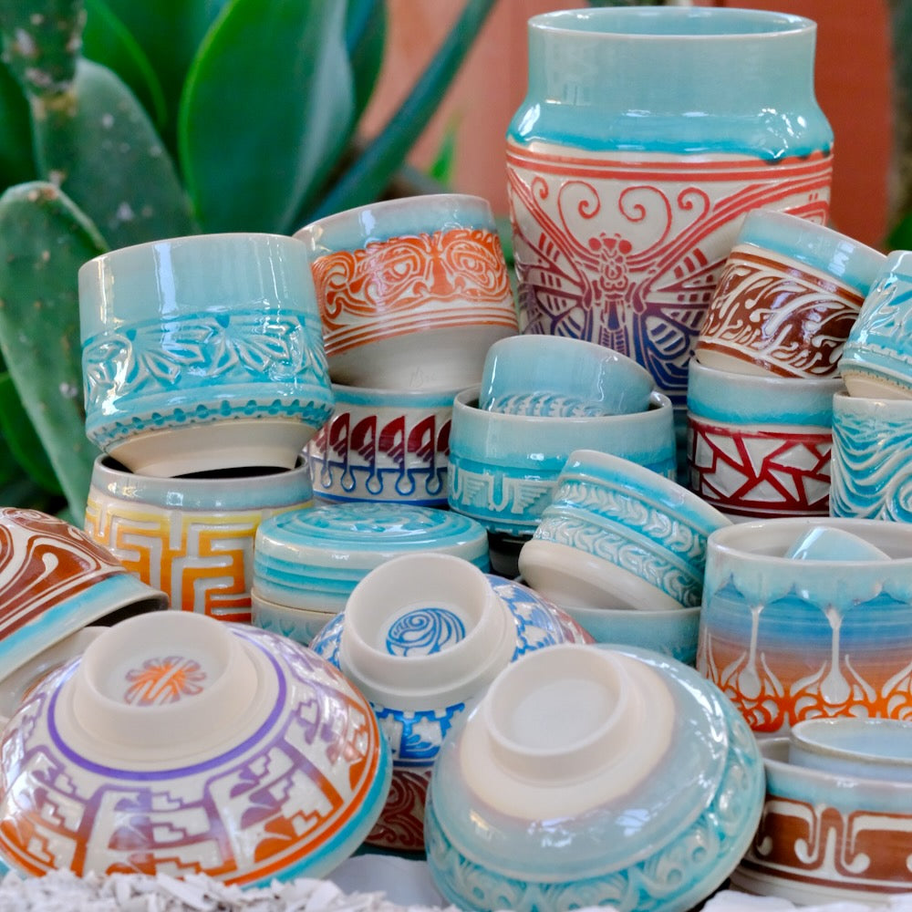 A collection of hand-painted ceramic bowls and small vessels stacked together, featuring traditional geometric and floral patterns in light blue, orange, red, purple, and brown.
