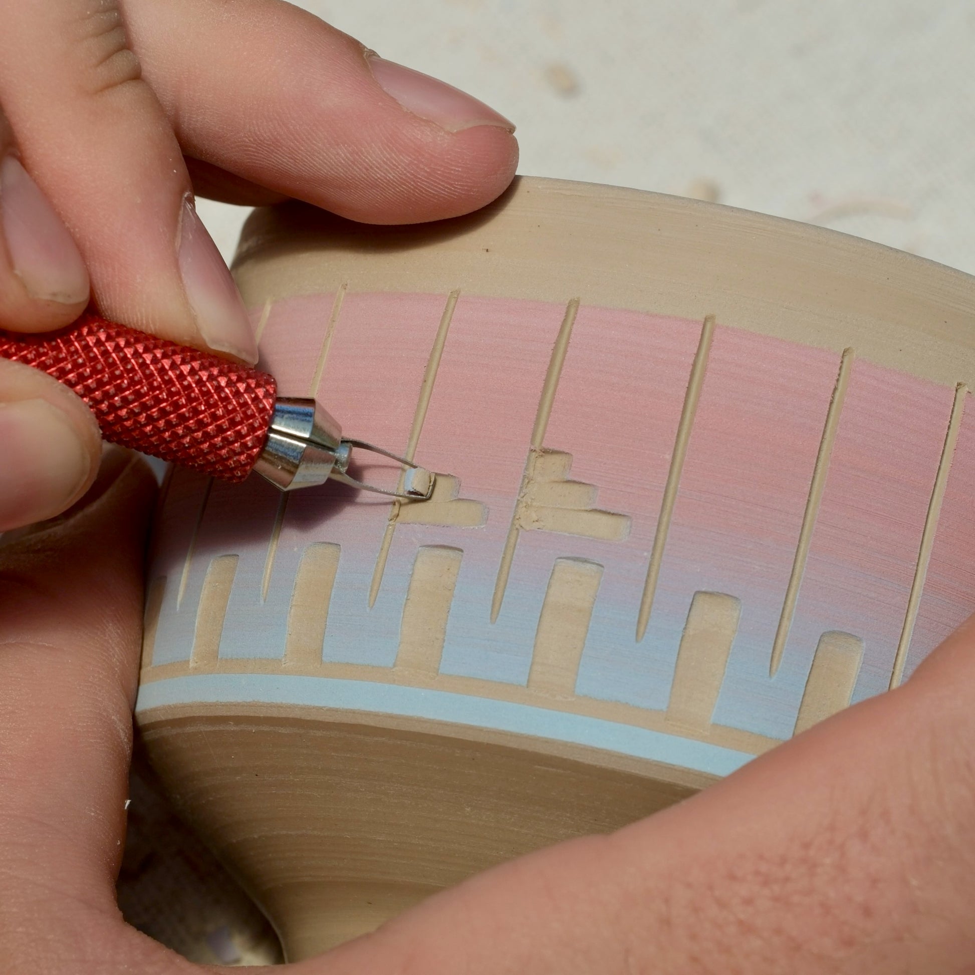 Close-up of a hand using a red-handled carving tool to etch lines into a light beige clay pottery piece with color on top.
