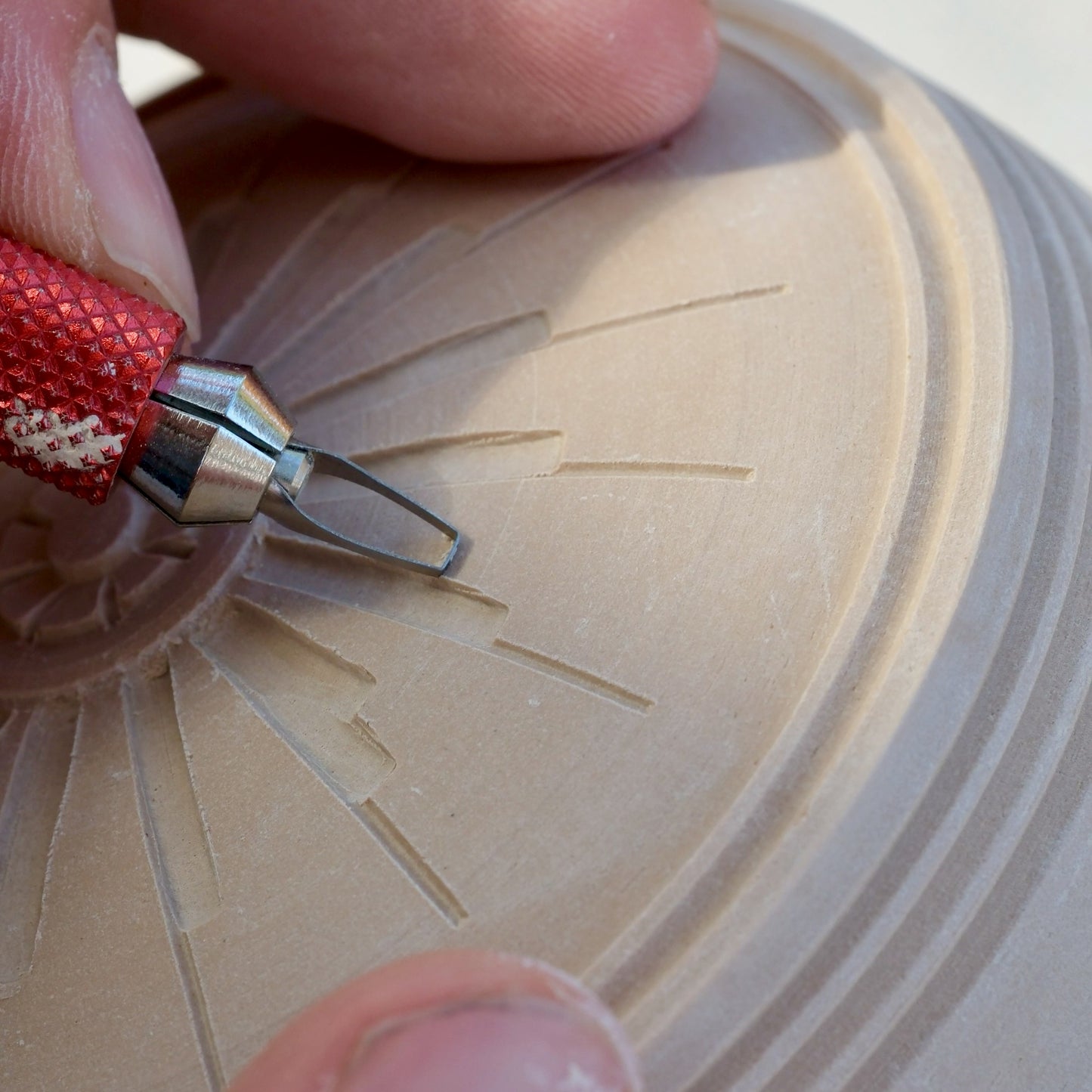 Close-up of a hand using a red-handled carving tool to etch radial lines into a light beige clay pottery piece.