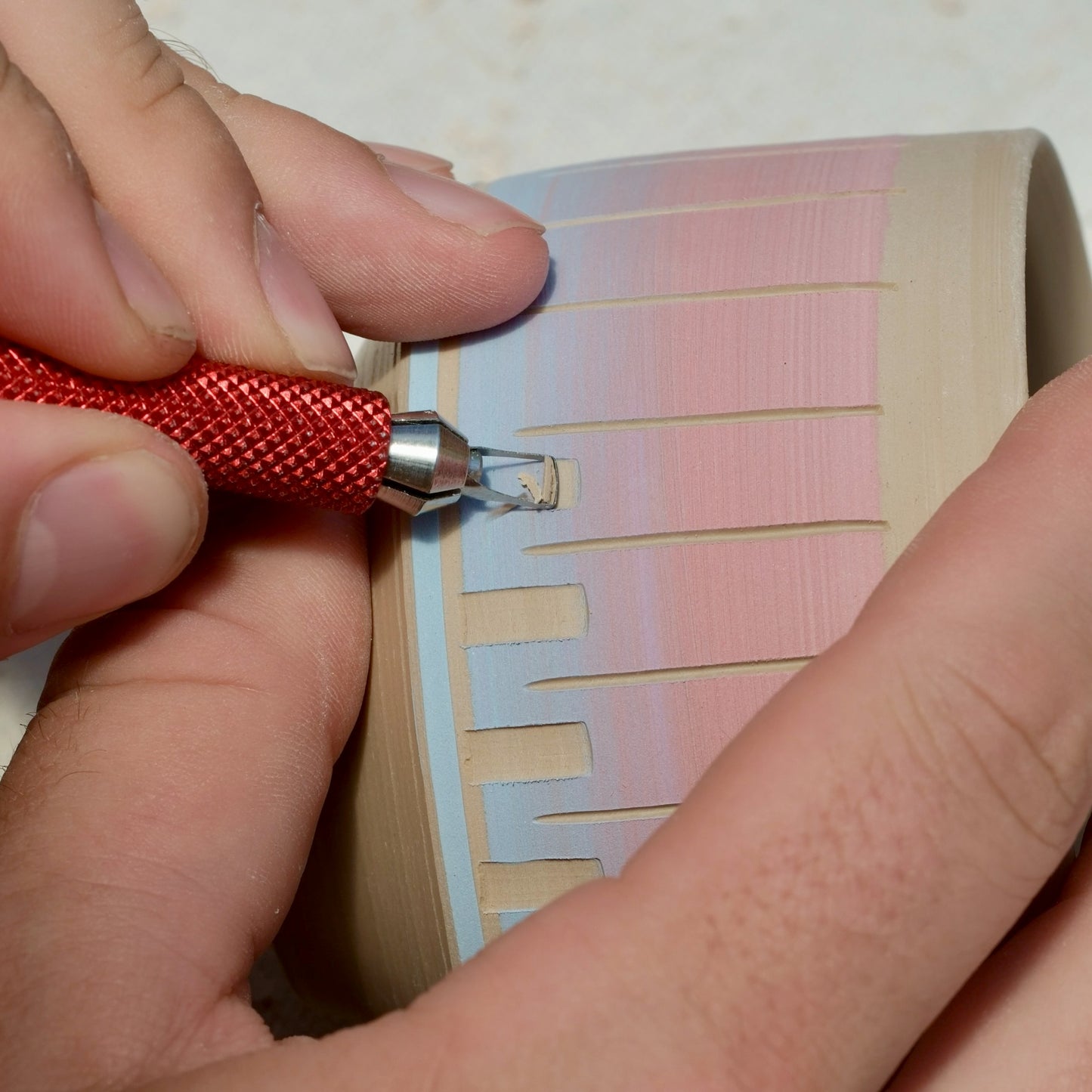 Close-up of a hand using a red-handled carving tool to etch lines into a light beige clay pottery piece with color on top.