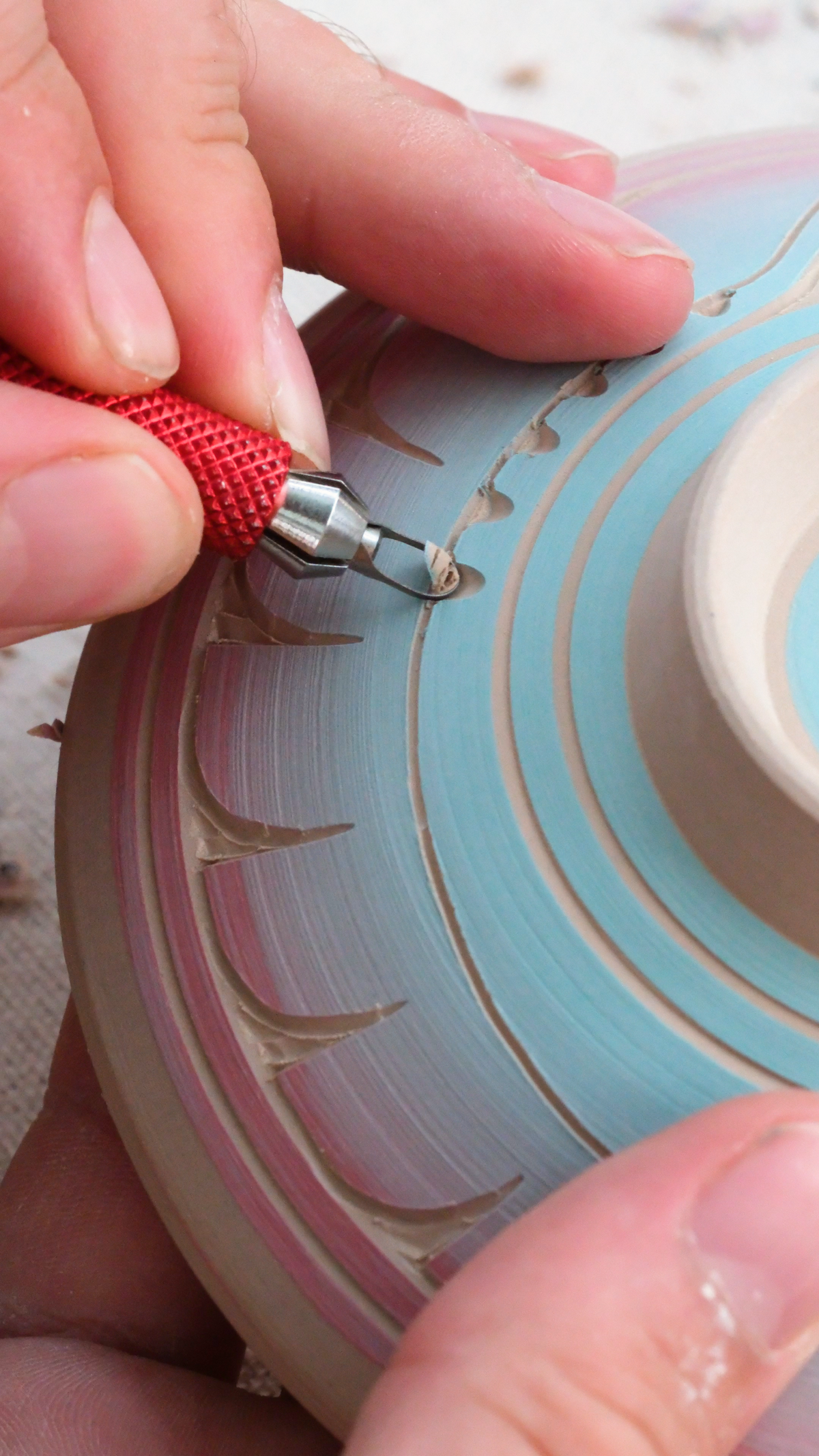 Close-up of a ceramic potter's wheel in progress, showing a hand using a red-handled ceramic carving tool to refine the textured, ridged surface of a handmade clay plate with concentric patterns in light blue, pink, and beige.