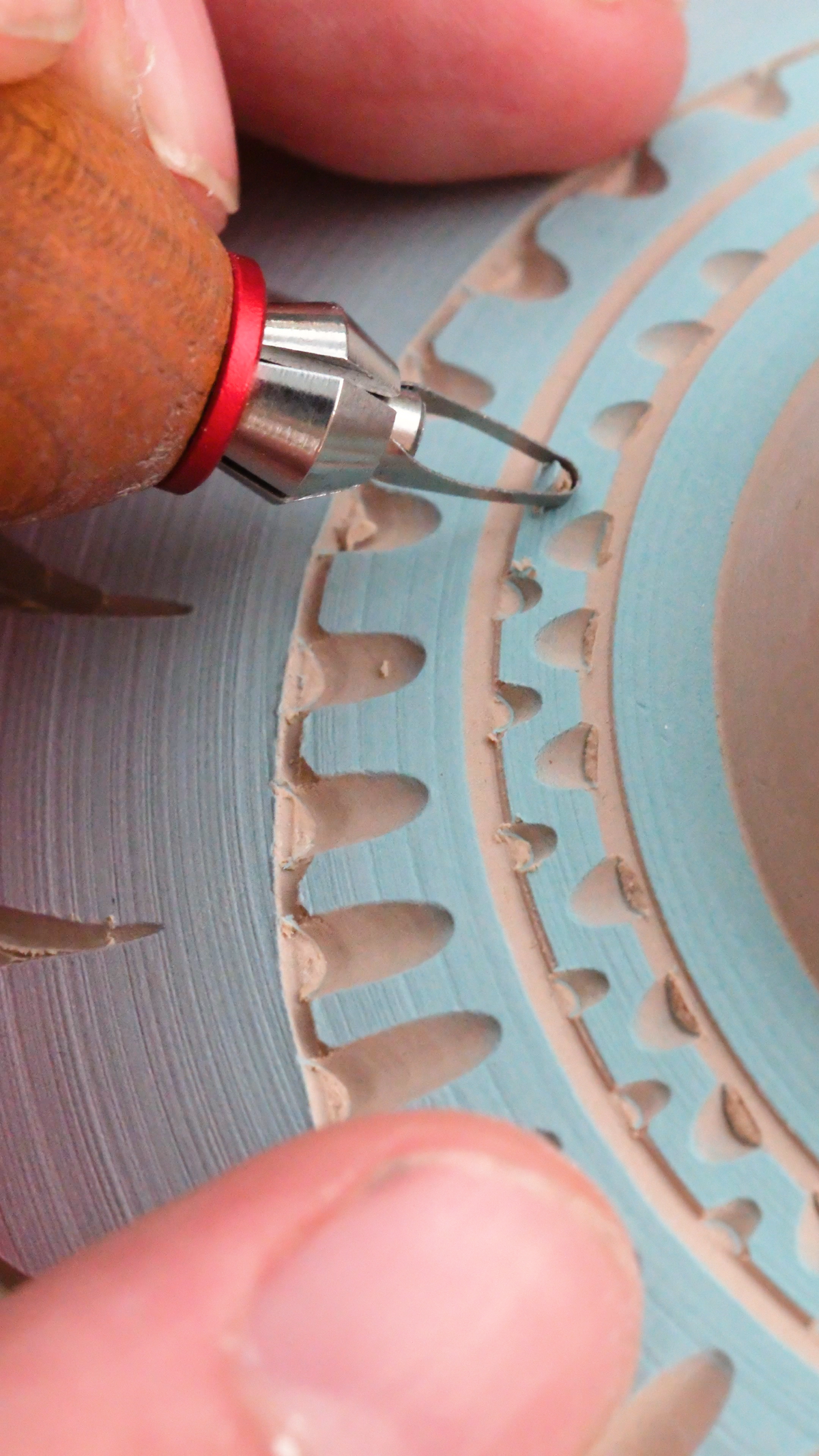 Close-up of a ceramic sculptor using a hand tool with a wooden handle and red collar to carve details into a light blue and beige ceramic wheel.