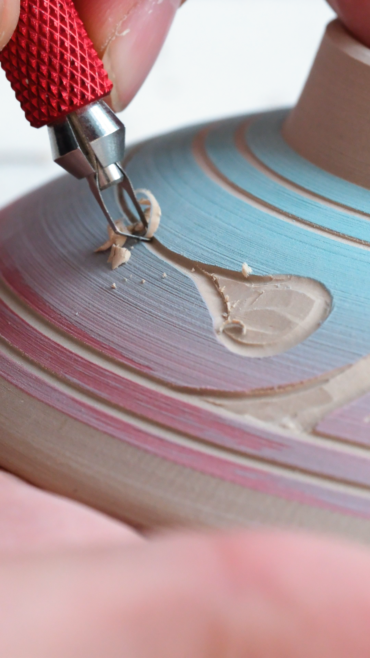Close-up of a ceramic pot being shaped on a wheel, with a hand holding a red-handled pottery tool and beige clay dust visible in the workspace.