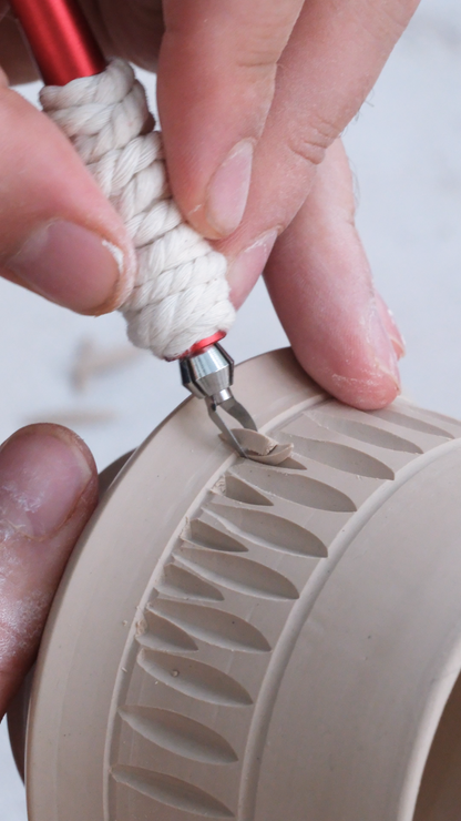Close-up of a hand using a small carving tool wrapped in white cord to shape a beige ceramic vessel with a ribbed pattern.