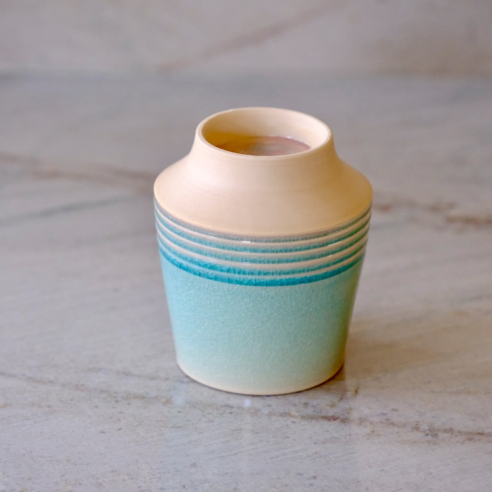 A small ceramic bud vase with a cream-colored top and a light blue-green glazed body featuring horizontal ridges, set on a light marble surface.