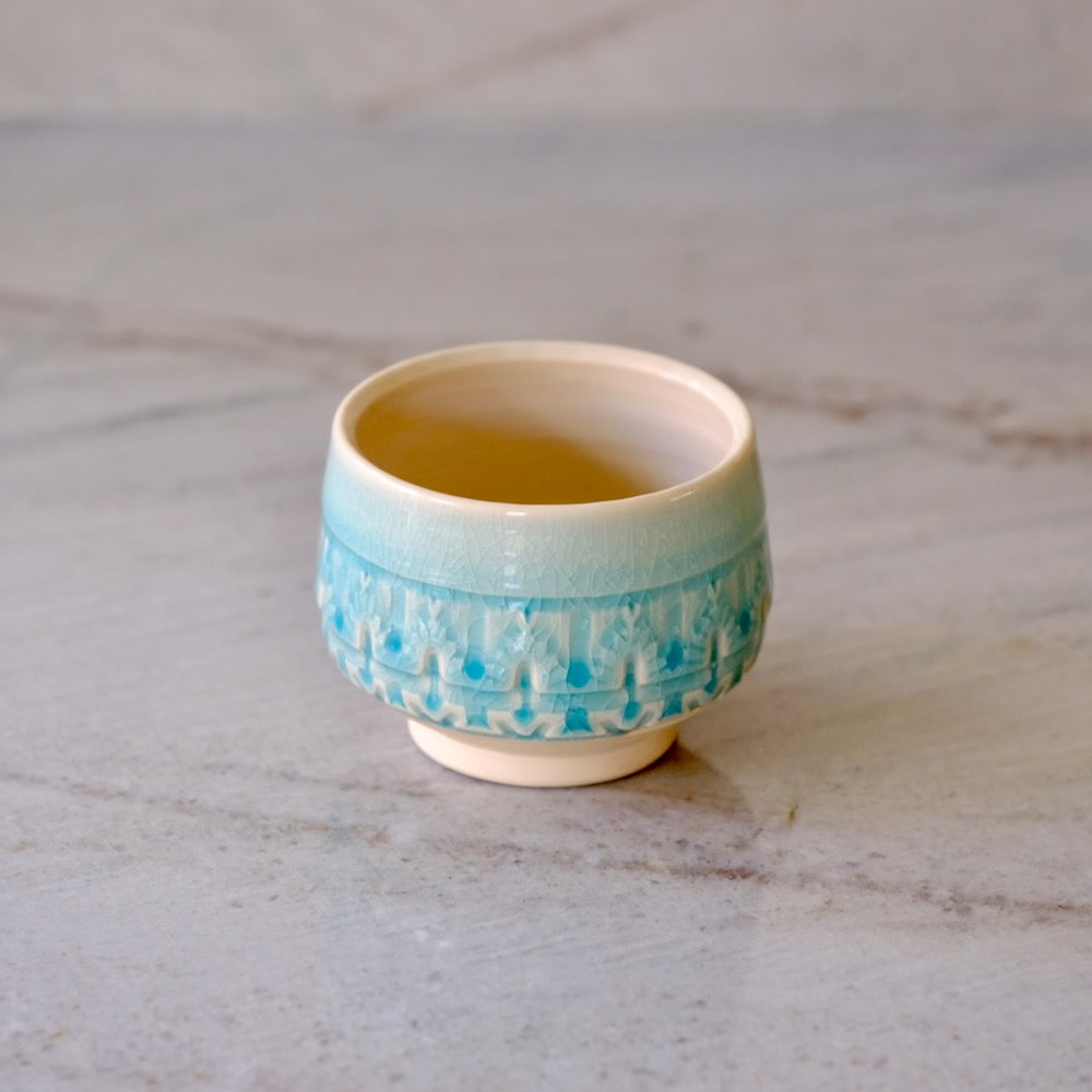 A small ceramic cup with a textured, ribbed turquoise blue band around the lower half and a warm cream interior, set on a light gray marble surface.