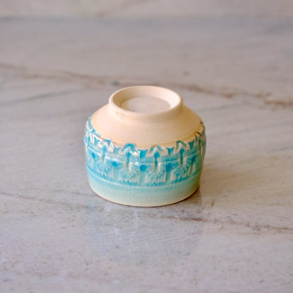 A small handmade ceramic cup with a cream-colored interior and exterior, featuring a textured turquoise blue pattern around the middle, resting on a light gray marble surface.