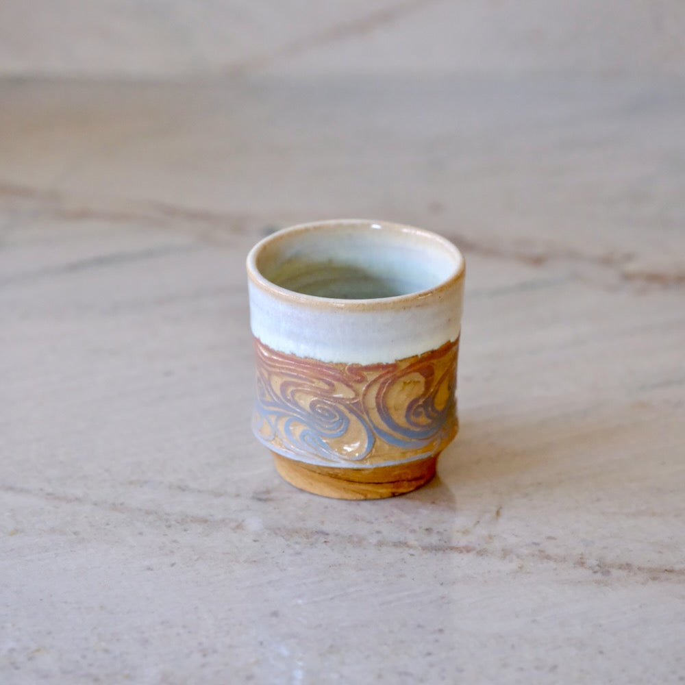 A small handmade ceramic cup with a light blue-green glazed interior, a white upper band, and a lower band featuring a marbled swirl pattern in gold, brown, and muted blue, set on a light stone surface.