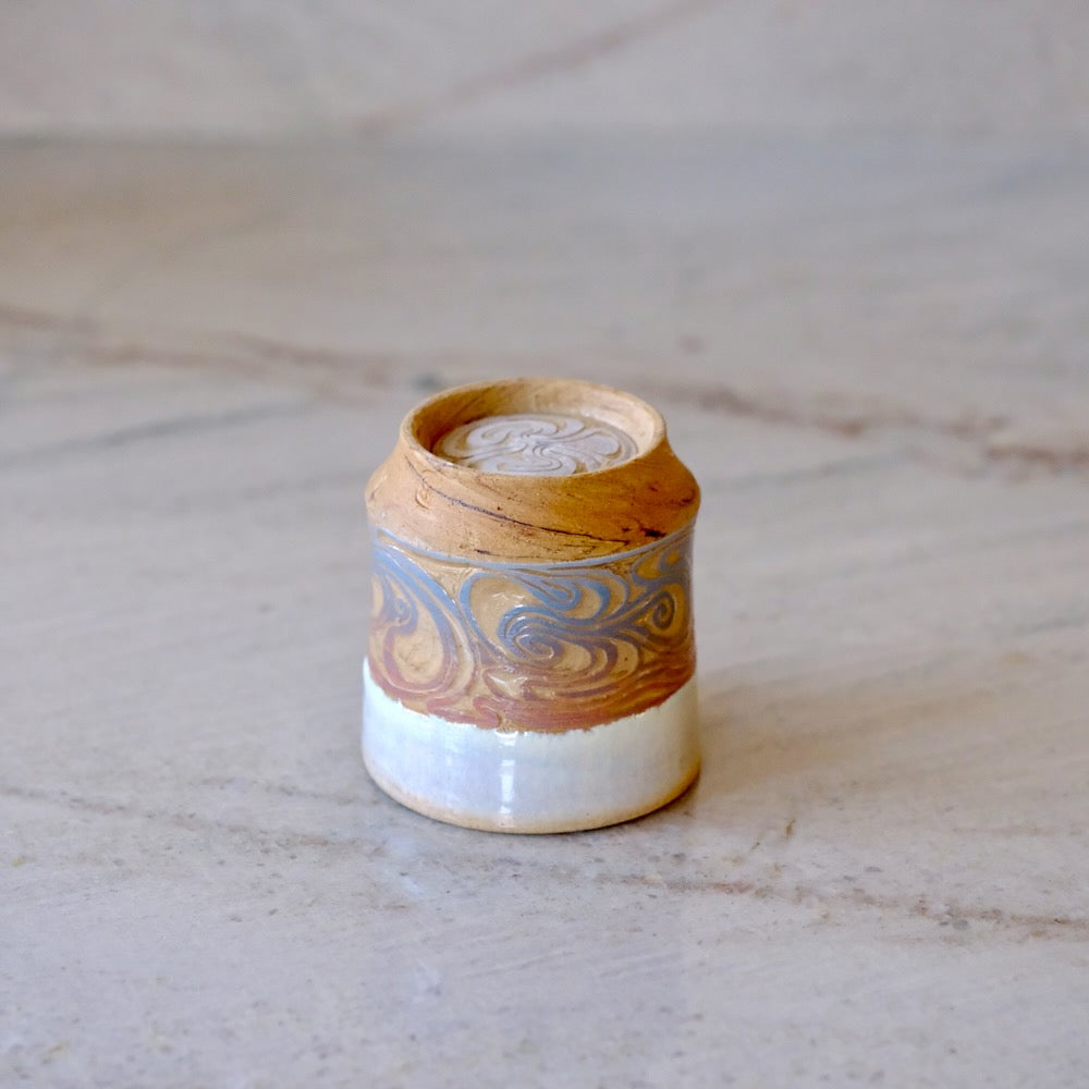 A small handmade ceramic container with a lid, featuring a swirled pattern in brown, gold, and blue-gray on a white glazed base, set on a light marble surface.
