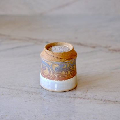 A small handmade ceramic container with a lid, featuring a swirled pattern in brown, gold, and blue-gray on a white glazed base, set on a light marble surface.