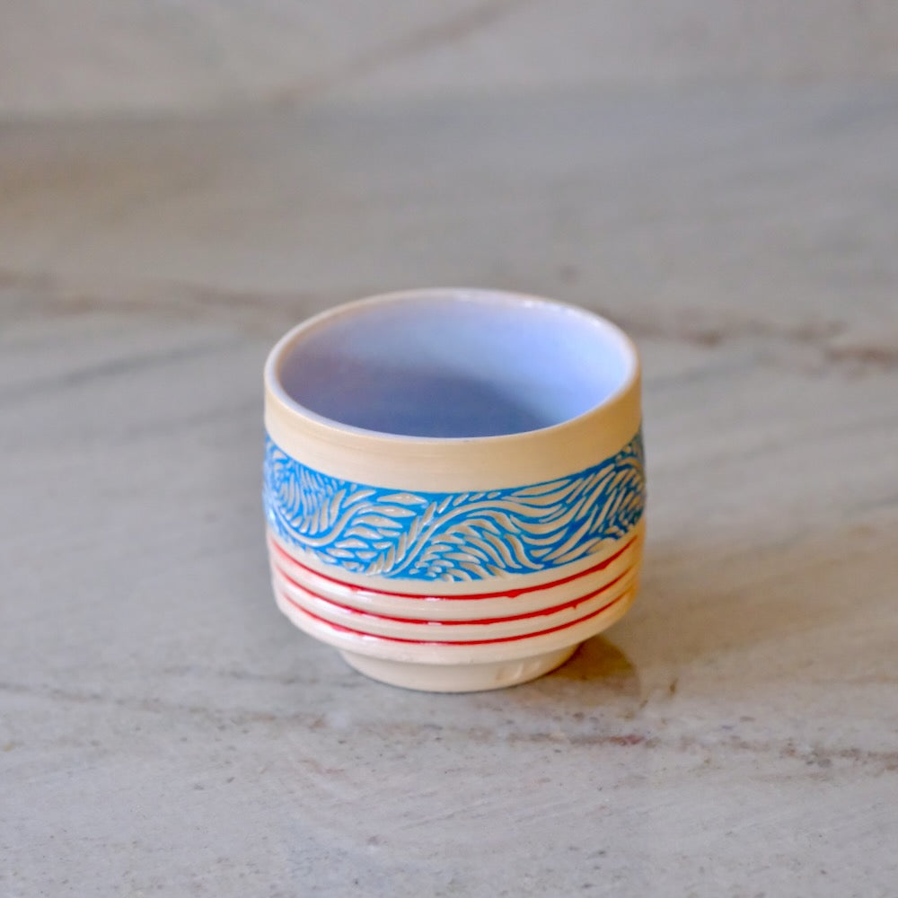 A small ceramic cup with a cream-colored exterior, featuring a blue wave pattern band and red horizontal stripes, set against a neutral gray stone background.