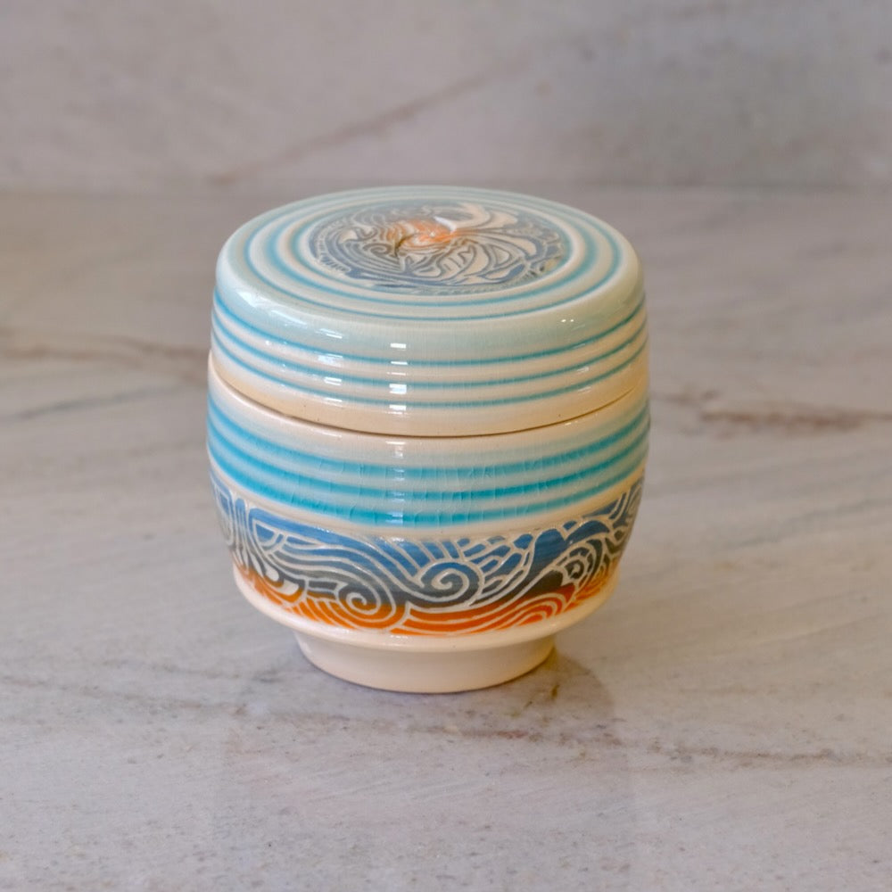 A small round ceramic lidded container with a cream-colored glaze, decorated with horizontal turquoise blue stripes and blue wave patterns, accented with orange near the base, set on a light marble surface.