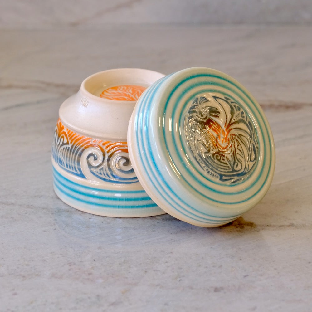 A small round ceramic lidded container with a matching lid leaning against the open base, shown on a light stone surface. The ceramic is off-white with turquoise-blue concentric stripes, blue wave motifs, and orange decorative accents, plus a painted rooster design on the lid.
