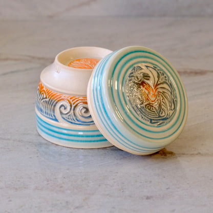 A small round ceramic lidded container with a matching lid leaning against the open base, shown on a light stone surface. The ceramic is off-white with turquoise-blue concentric stripes, blue wave motifs, and orange decorative accents, plus a painted rooster design on the lid.