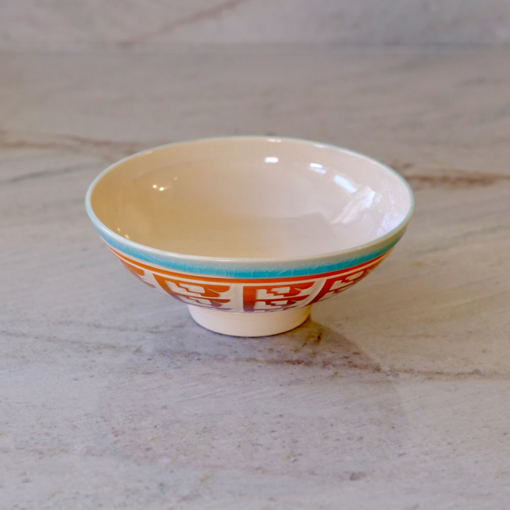 A small ceramic bowl with a cream-colored interior, a turquoise rim stripe, and orange Chinese characters printed on the exterior, set on a light marble surface.