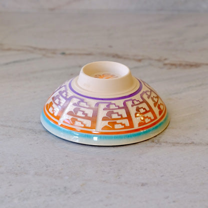 A small ceramic bowl with a white base, decorated with a geometric pattern in orange, purple, and turquoise blue, photographed on a light gray marble surface.