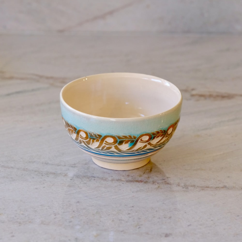 A small ceramic bowl with a light blue exterior, decorated with a gold and brown floral pattern and a cream-colored interior, resting on a light marble surface.