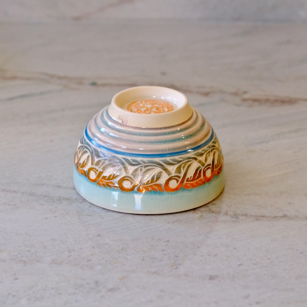 A small ceramic bowl with a colorful hand-painted pattern, featuring blue, orange, and white designs on a light beige base, photographed on a light gray stone surface.