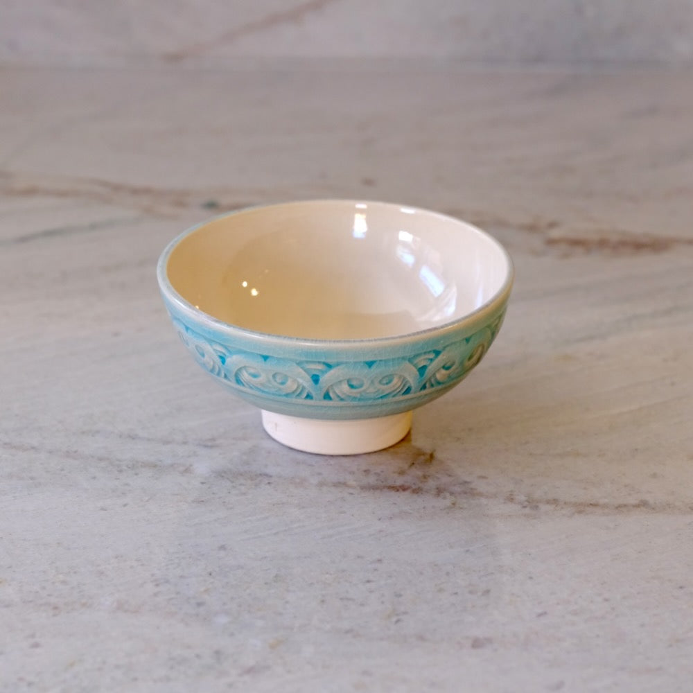 A small ceramic bowl with a cream-colored interior and exterior base, decorated with a turquoise blue patterned band around the middle, set on a light marble surface.