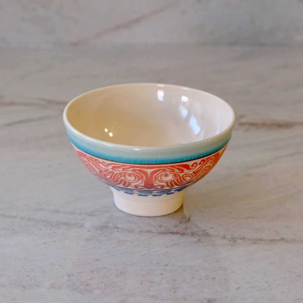 A small ceramic bowl with a cream-colored interior, a turquoise rim, and a red and blue patterned exterior, set on a light marble surface.