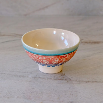 A small ceramic bowl with a cream-colored interior, a turquoise rim, and a red and blue patterned exterior, set on a light marble surface.