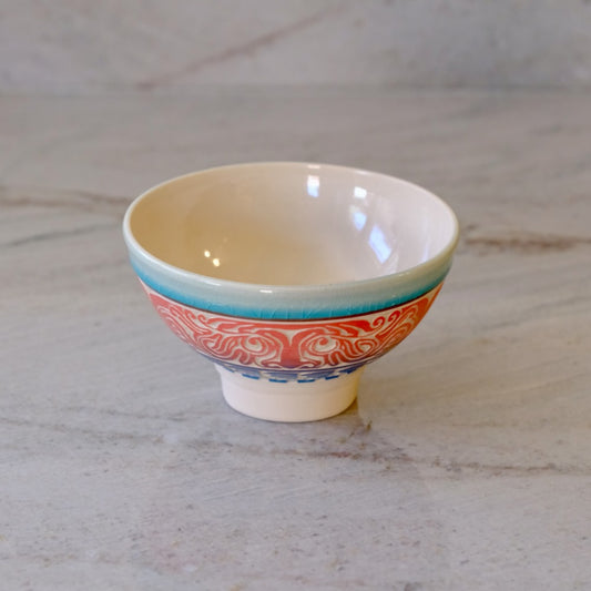 A small ceramic bowl with a cream-colored interior, a turquoise rim, and a red and blue patterned exterior, set on a light marble surface.