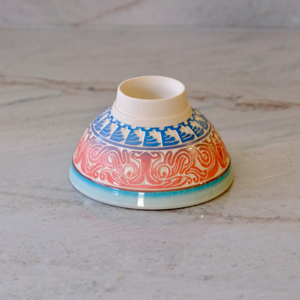 A small ceramic bowl with a wide rim, decorated with a blue geometric pattern and a red-orange scrollwork design, set on a light gray marble surface.
