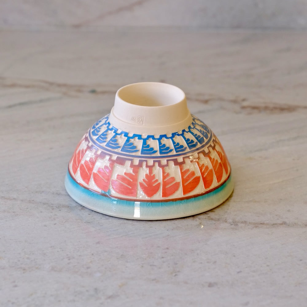 A small ceramic bowl with a hand-painted pattern of red leaves and blue geometric accents, shown upside down on a light gray marble surface.