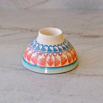 A small ceramic bowl with a hand-painted pattern of red leaves and blue geometric accents, shown upside down on a light gray marble surface.