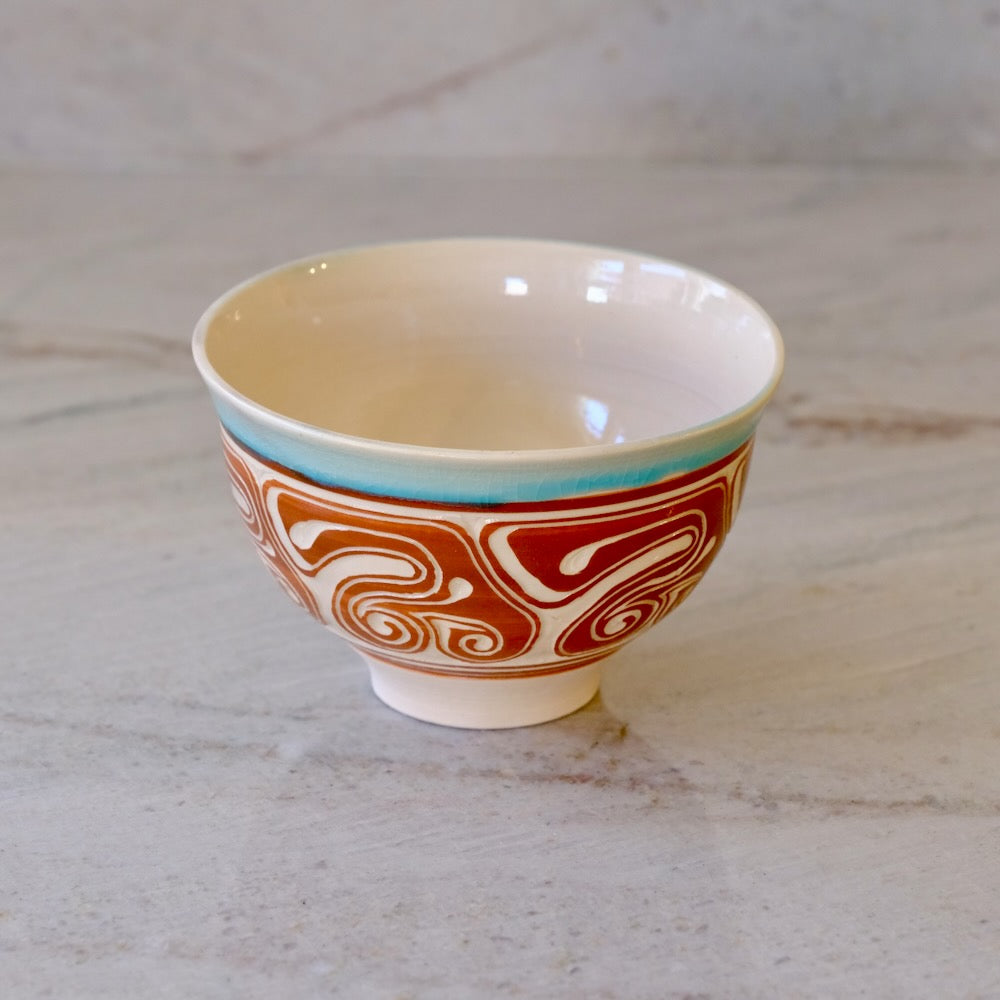 A ceramic bowl with a cream-colored interior, a turquoise band near the rim, and a red and white abstract wave pattern on the exterior, set on a light marble surface.