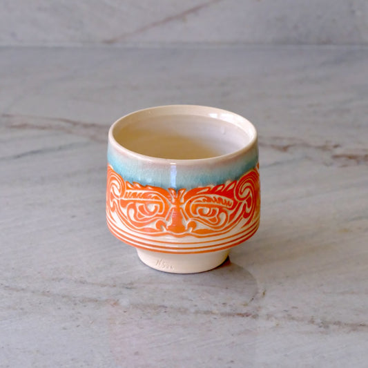 A small ceramic cup with a cream interior and exterior, featuring a turquoise band near the rim and a prominent orange and white stylized face pattern around the middle, set on a light gray marble surface.