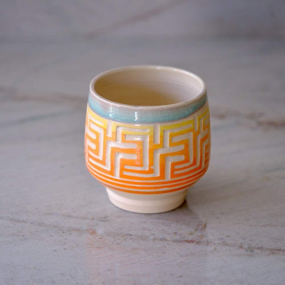 A small ceramic cup with a cream-colored interior and exterior, decorated with a yellow and orange geometric pattern and a light blue band near the rim, set on a light gray marble surface.