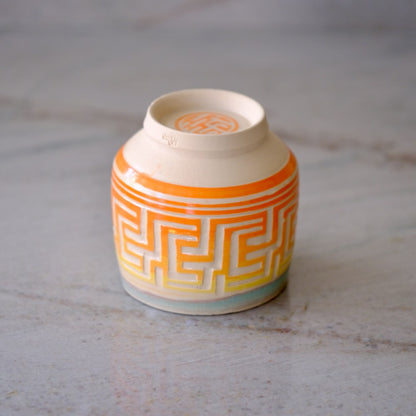 A small ceramic cup with a cream-colored interior and exterior, decorated with a geometric maze pattern in orange and yellow, resting on a light gray marble surface.