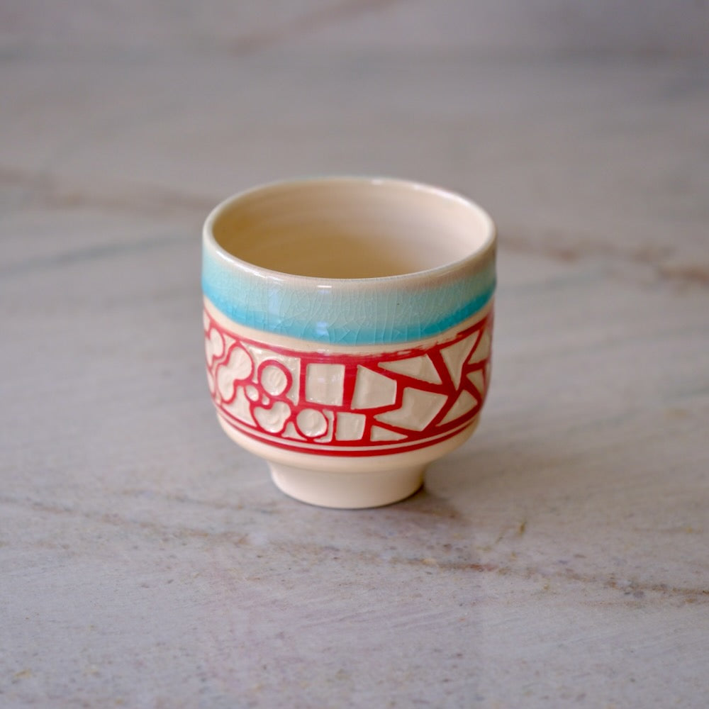 A small ceramic cup with a cream-colored glaze, featuring a turquoise blue band near the rim and a red geometric pattern around the middle, set on a light gray marble surface.