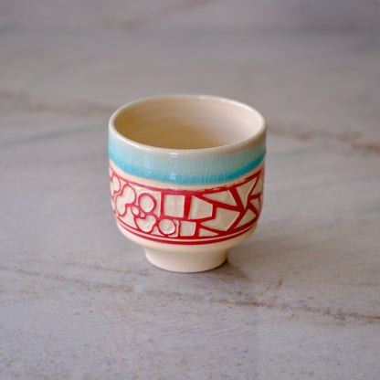 A small ceramic cup with a cream-colored glaze, featuring a turquoise blue band near the rim and a red geometric pattern around the middle, set on a light gray marble surface.