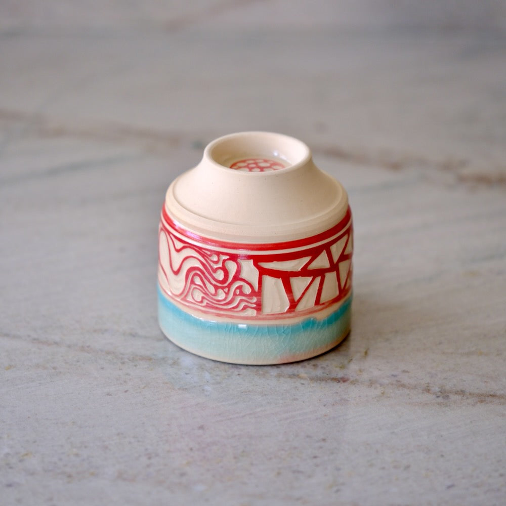 A small ceramic cup with a cream-colored body, decorated with red geometric and wave patterns and a light blue glazed base, set on a light gray marble surface.