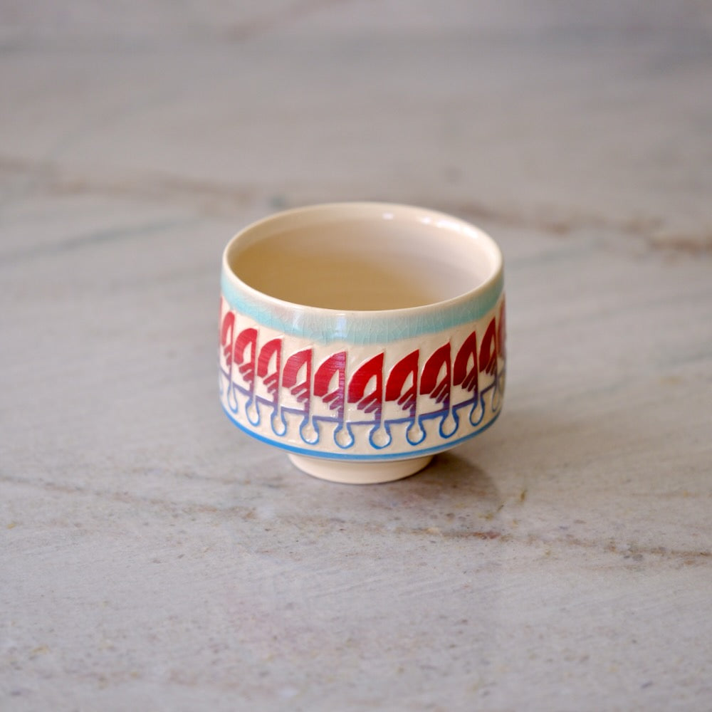 A small ceramic cup with a cream-colored interior and exterior, decorated with a repeating red and blue pattern around the lower half, set on a light gray marble surface.