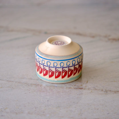 A small handmade ceramic cup with a lid, featuring a hand-painted pattern of red teardrops and blue loops on a cream-colored base, set on a light gray stone surface.