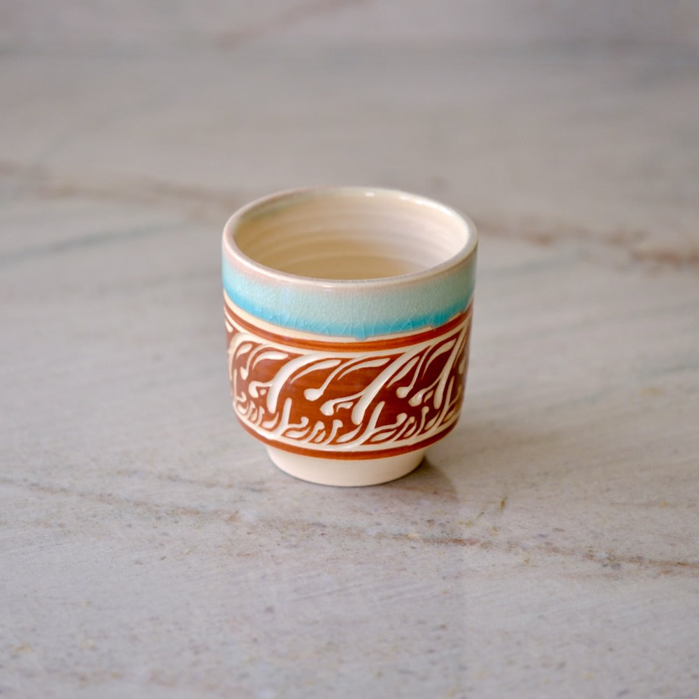 A small ceramic cup with a cream-colored interior and exterior, decorated with a turquoise blue band near the rim and a brown and white patterned wrap around the middle, set on a light marble surface.