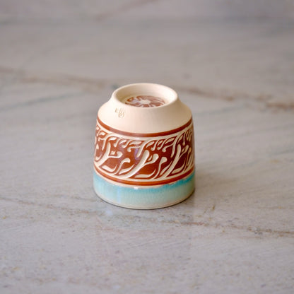 A small ceramic cup with a cream-colored body, featuring a brown band decorated with white Arabic calligraphy and a light blue glazed base, set on a light gray marble surface.