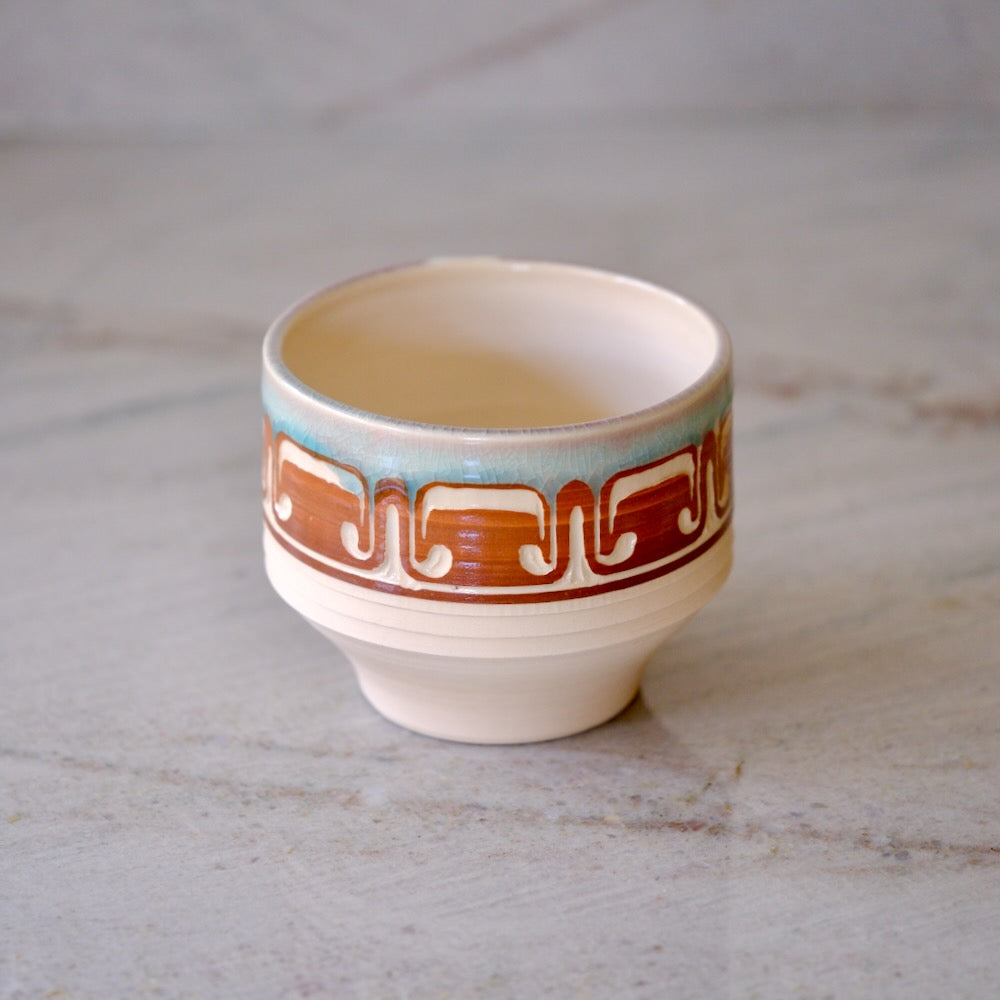 A small ceramic cup with a cream-colored body, featuring a decorative band with a brown geometric pattern and a light blue accent above it, set on a light gray marble surface.