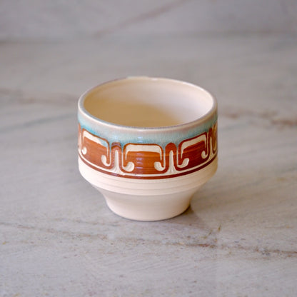 A small ceramic cup with a cream-colored body, featuring a decorative band with a brown geometric pattern and a light blue accent above it, set on a light gray marble surface.