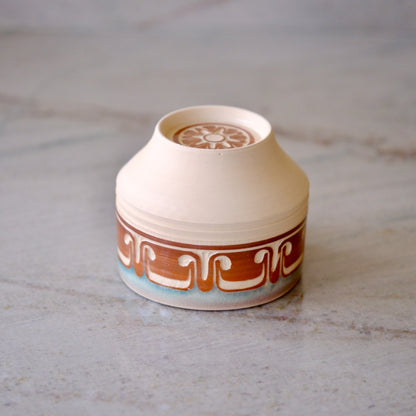 A small handmade ceramic container with a conical lid, featuring a cream-colored body, a brown geometric pattern band, and a light blue glaze at the base, set on a light gray stone surface.
