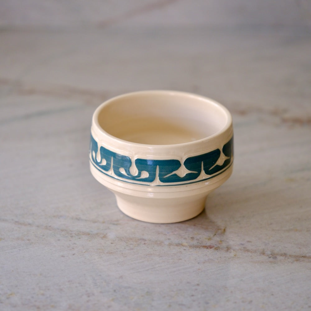 A small cream-colored ceramic cup with a teal blue decorative band featuring a repeating wave-like pattern, set on a light gray marble surface.