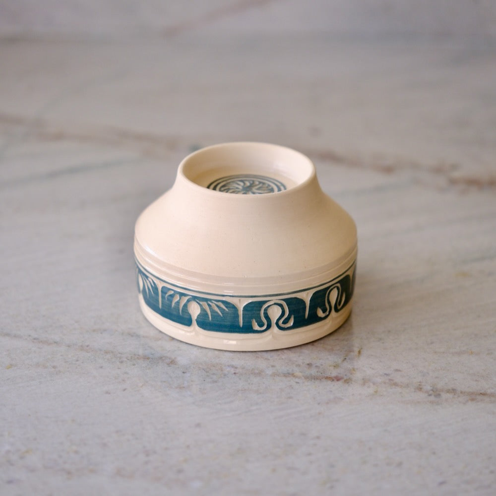 A small cream-colored ceramic cup with a dark teal blue wave pattern band around the lower half, shown on a light marble surface.